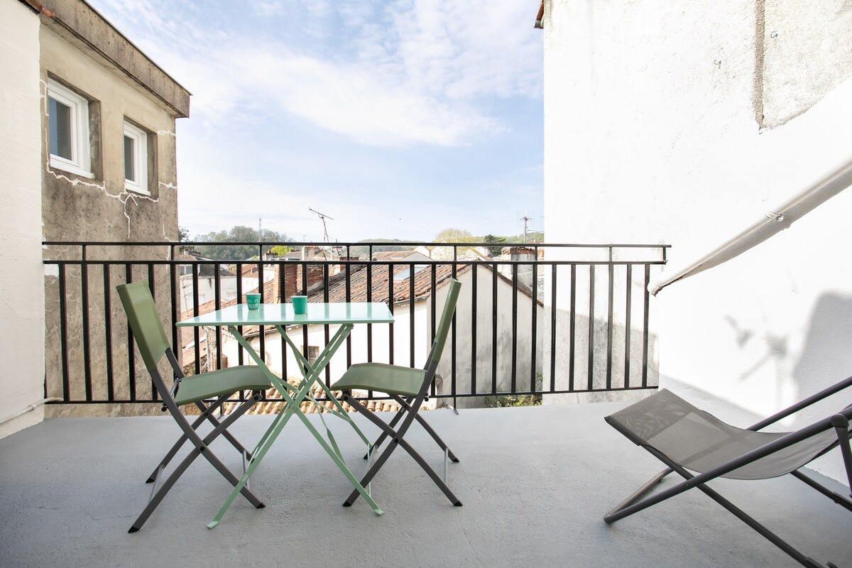 A balcony is presented with a small glass table surrounded by two green chairs. A reclined chair is positioned nearby, offering a relaxed seating option. The view includes neighboring rooftops and clear skies in the background, enhancing the outdoor experience.