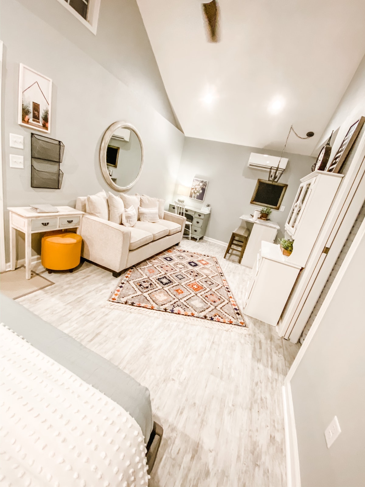 A studio apartment features a cozy couch and a compact desk area. A decorative rug adds warmth to the light-colored floor. A round mirror reflects the softly lit space, while an air conditioning unit is positioned for comfort. Walls are painted in neutral tones, enhancing the room's brightness.