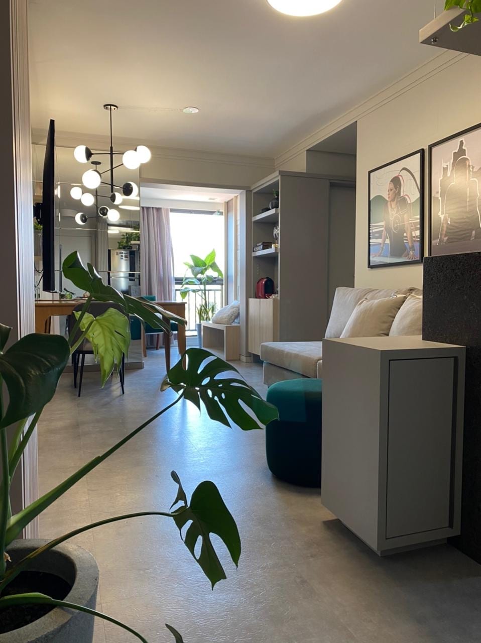 A well-lit, modern living area features a comfortable seating arrangement with plush cushions. A large window offers a view of greenery outside, while a chic chandelier hangs overhead. Decorative shelves provide functional storage, and a potted plant adds a touch of nature to the space.