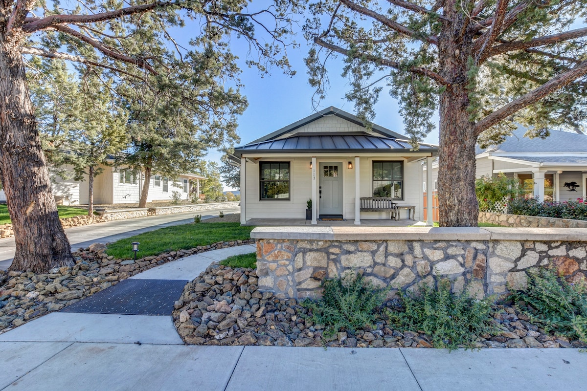 A charming bungalow exterior is framed by tall trees, with a stone wall and pathway leading to the entrance. The welcoming porch includes a bench, while flower beds add natural greenery to the inviting façade.