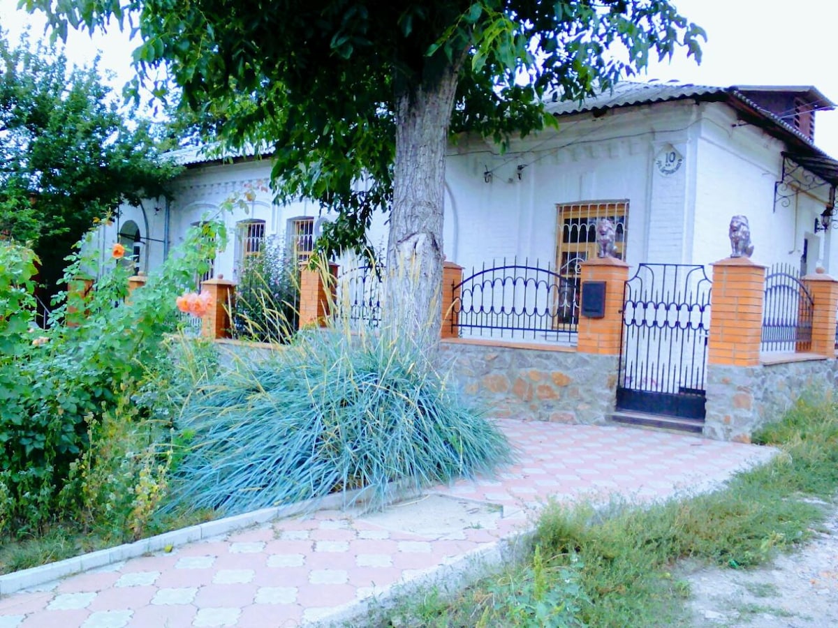 A welcoming entrance is framed by a lush green landscape, featuring a pathway made of patterned tiles leading to a decorative wrought-iron gate. Natural vegetation surrounds the property, enhancing the tranquil atmosphere of the exterior.