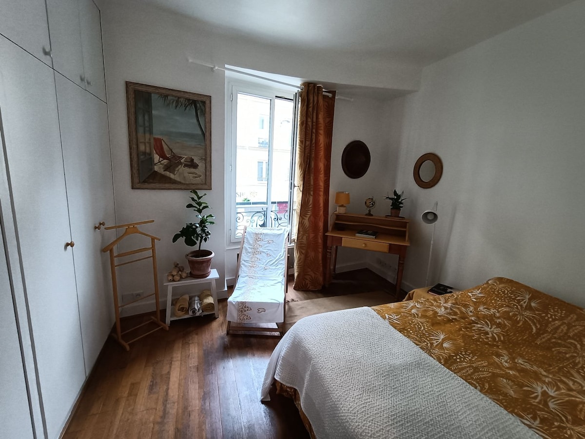 A bright bedroom features a spacious bed with a patterned blanket and light streaming in through a large window. A wooden desk stands next to the window, accompanied by a small plant. Decorative wall art and a clothing rack are visible, adding to the room's character.