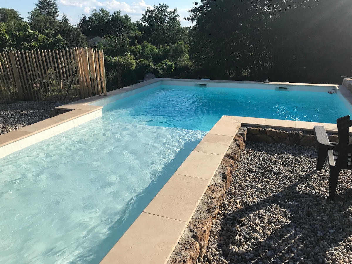 A tranquil swimming pool is shown, featuring two distinct sections with clear blue water. Surrounding the pool are well-maintained pebbled areas and a wooden fence that offers privacy. A wooden chair is positioned nearby, inviting relaxation amid the serene landscape.