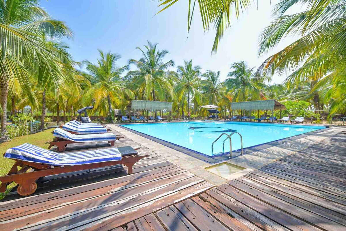 A spacious swimming pool is surrounded by palm trees, offering a serene atmosphere. Lounge chairs with striped cushions are positioned on a wooden deck, inviting relaxation. Sunlight reflects off the water's surface, highlighting the peaceful surroundings and clear blue sky.