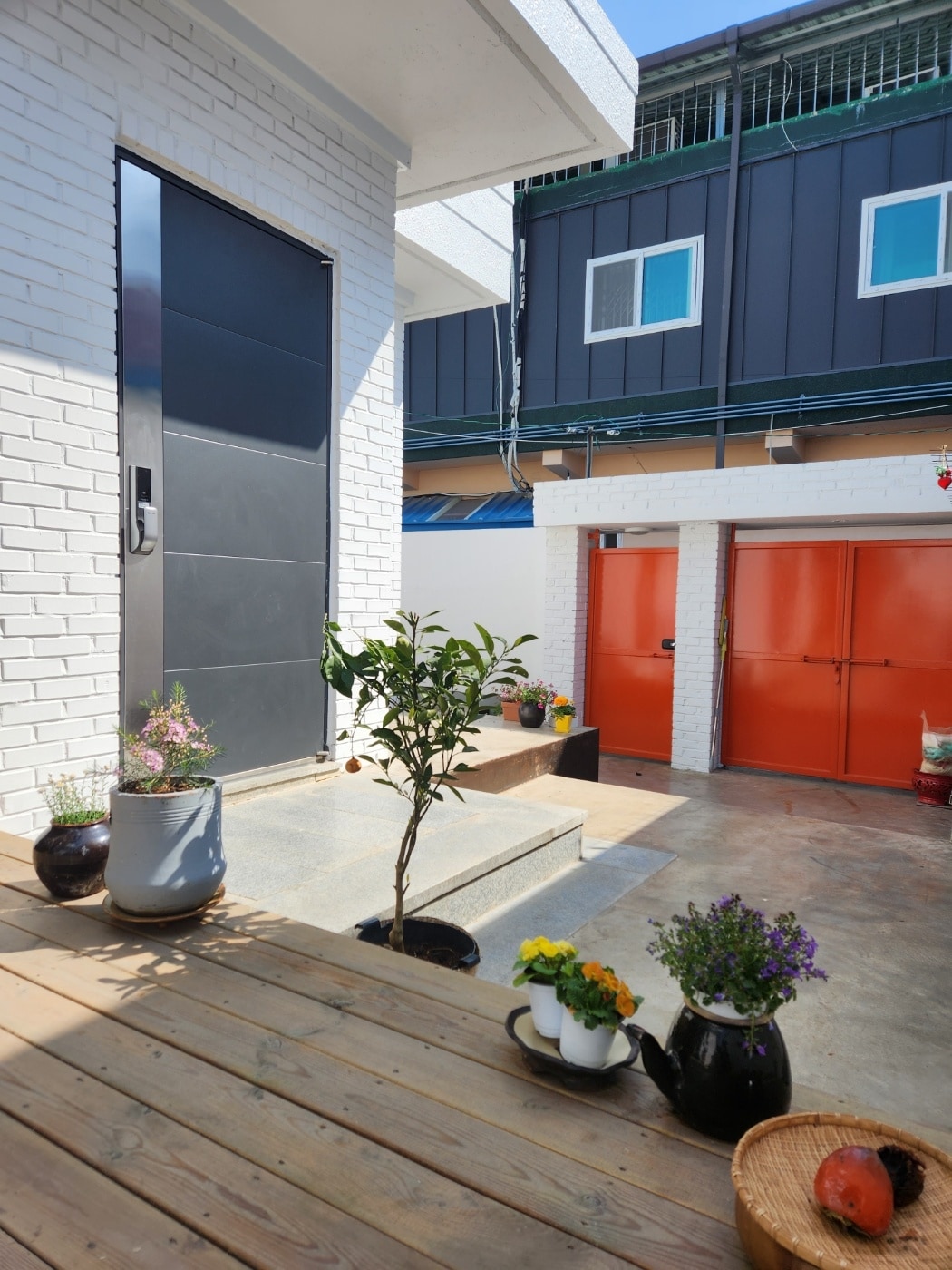 The entrance features a modern black door set against a white brick wall, flanked by potted plants. A small wooden deck leads to the entrance, with colorful flowers arranged in pots. A light-filled courtyard is present, alongside garage doors painted in bright orange.