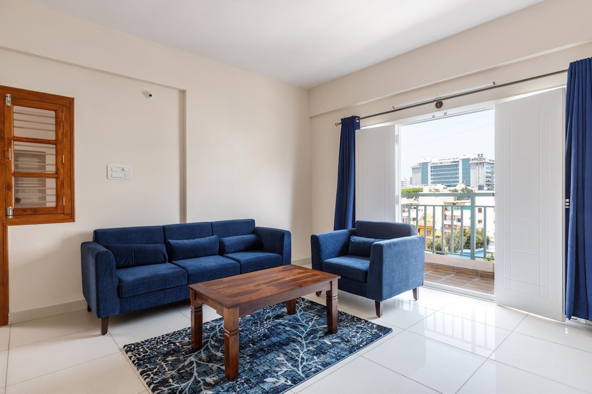 A bright living area features a blue sofa and armchair arranged around a wooden coffee table. A patterned rug lies underneath, while large windows are framed by blue curtains, allowing natural light to fill the space and offering views of the outside.