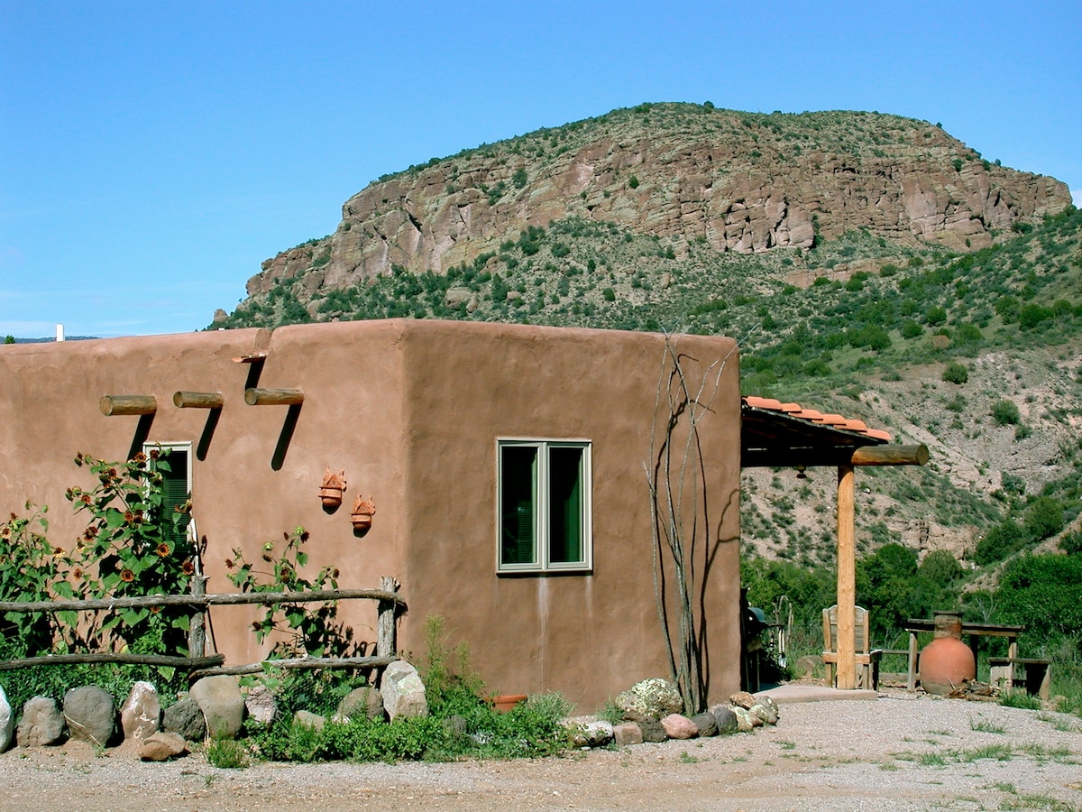 A single-story adobe casita is set against a backdrop of rugged hills. Natural materials are used in the construction, with green windows and a clay tile roof. Nearby, a shaded porch area is provided with rustic wooden support beams.