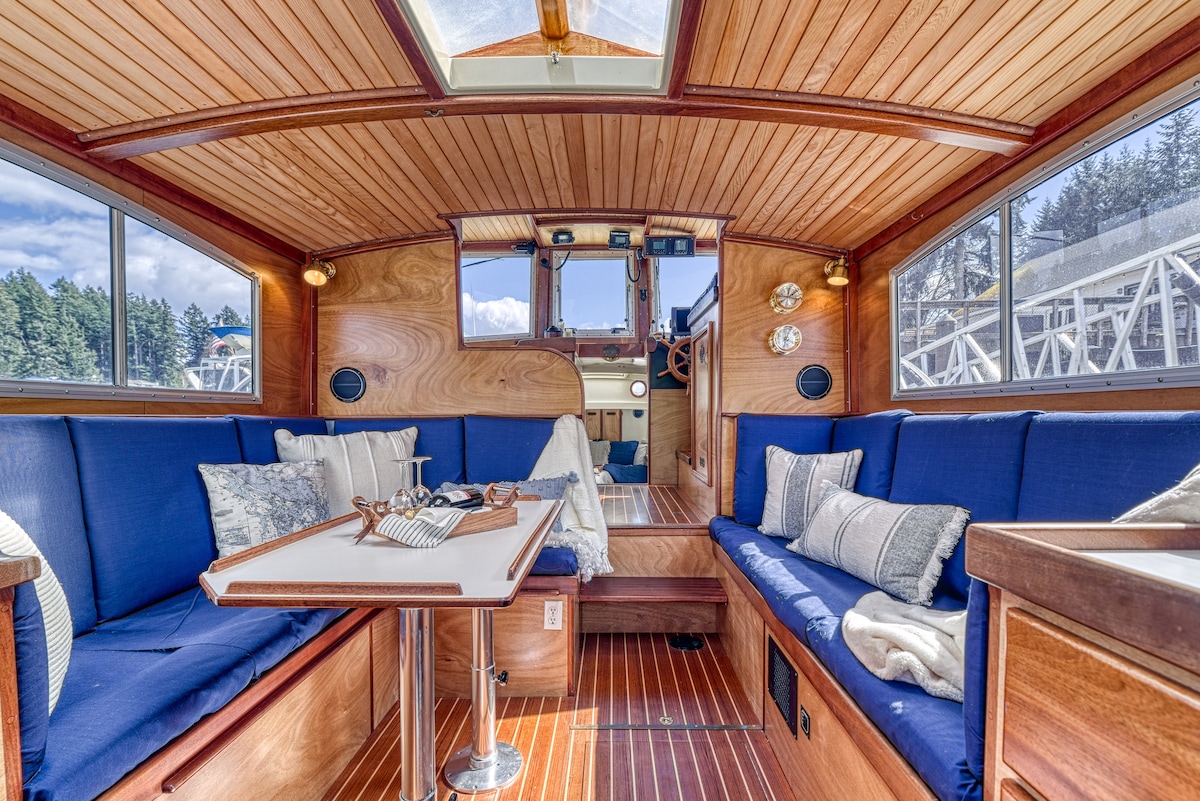 The interior seating area features blue upholstery with decorative pillows, creating a comfortable space. A central table is set for dining, surrounded by wood paneling and large windows that allow natural light to fill the boat. The helm is visible in the front.