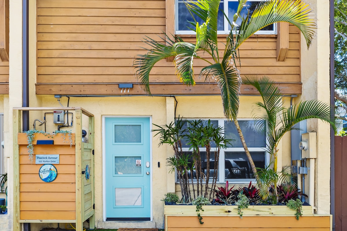The entrance of the townhouse is adorned with a light blue door and a decorative sign. Lush greenery, including palm trees and colorful plants, enhances the welcoming atmosphere. Large windows allow natural light to fill the space, highlighting the inviting exterior.