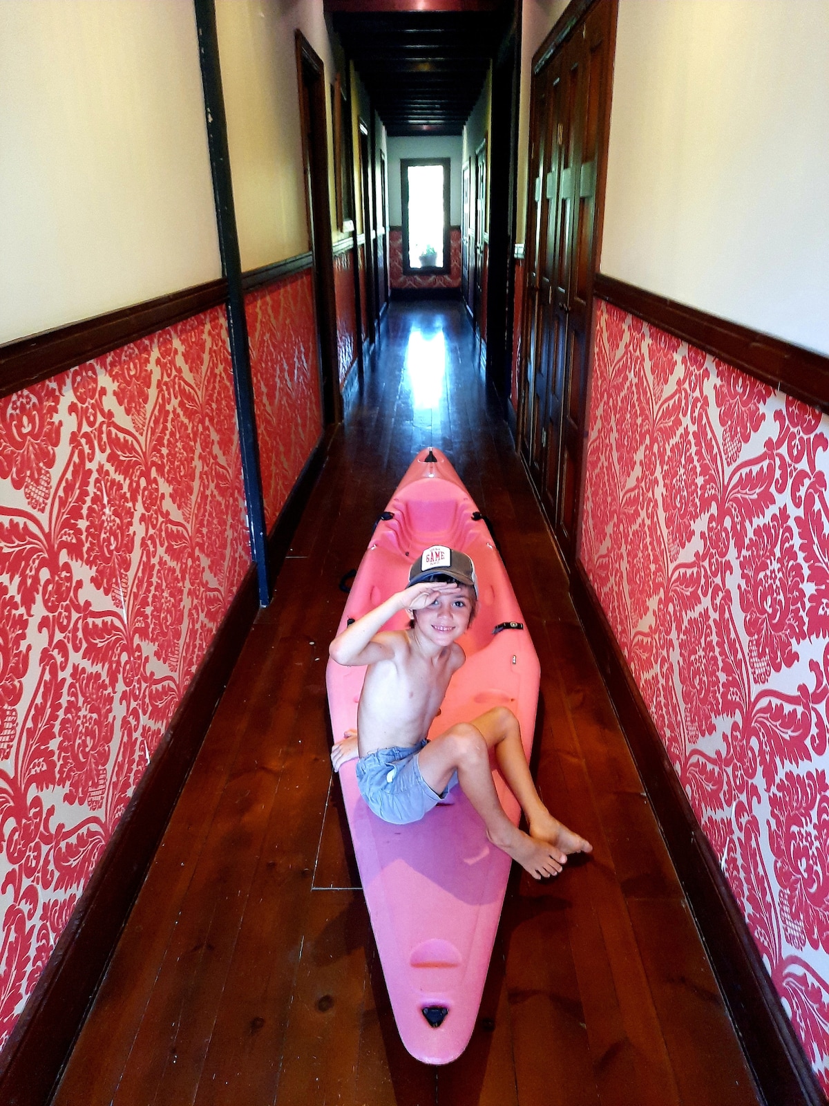 A colorful hallway features strikingly patterned wallpaper in shades of red. A smooth wooden floor runs the length of the corridor, illuminated by natural light entering from the far end. A pink kayak is positioned along the floor, with a child seated inside, adding a playful touch to the setting.