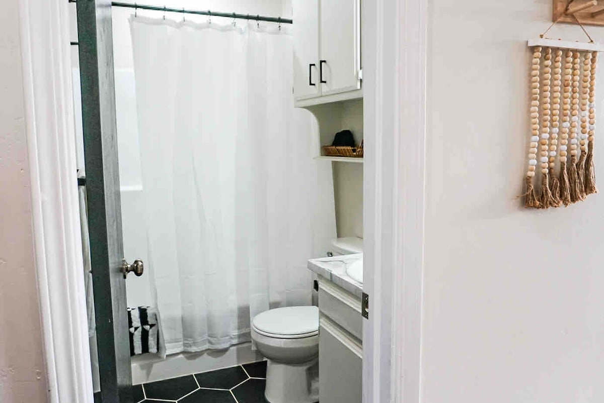 A bright bathroom is presented with a white shower curtain and hexagonal floor tiles. A white toilet is positioned next to a compact sink and cabinet. Natural light filters in from the frosted glass panel, creating a clean and simple atmosphere.