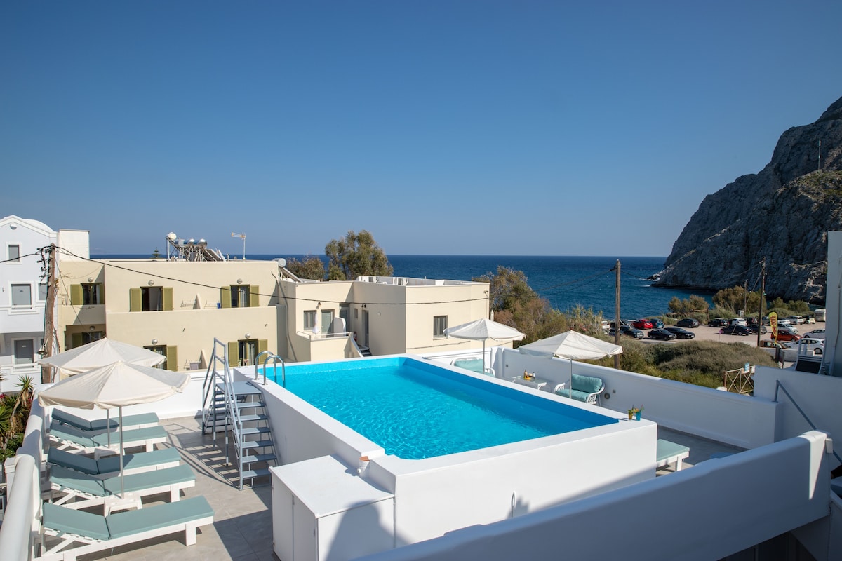 An outdoor pool is surrounded by lounge chairs and umbrellas, with a scenic view of the sea and rocky cliffs in the background. The space is bright and airy, reflecting a serene atmosphere for relaxation.