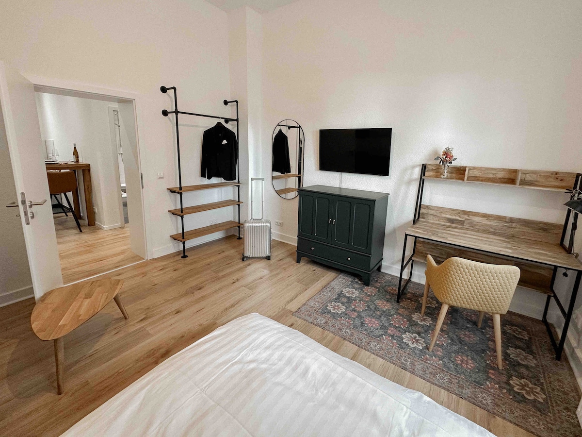 An open room features a comfortable sleeping area with white bedding. A wooden desk and chair are situated near a television. A clothing rack and mirror provide functionality, while a small table and rug add warmth. The room is well-lit with natural light.