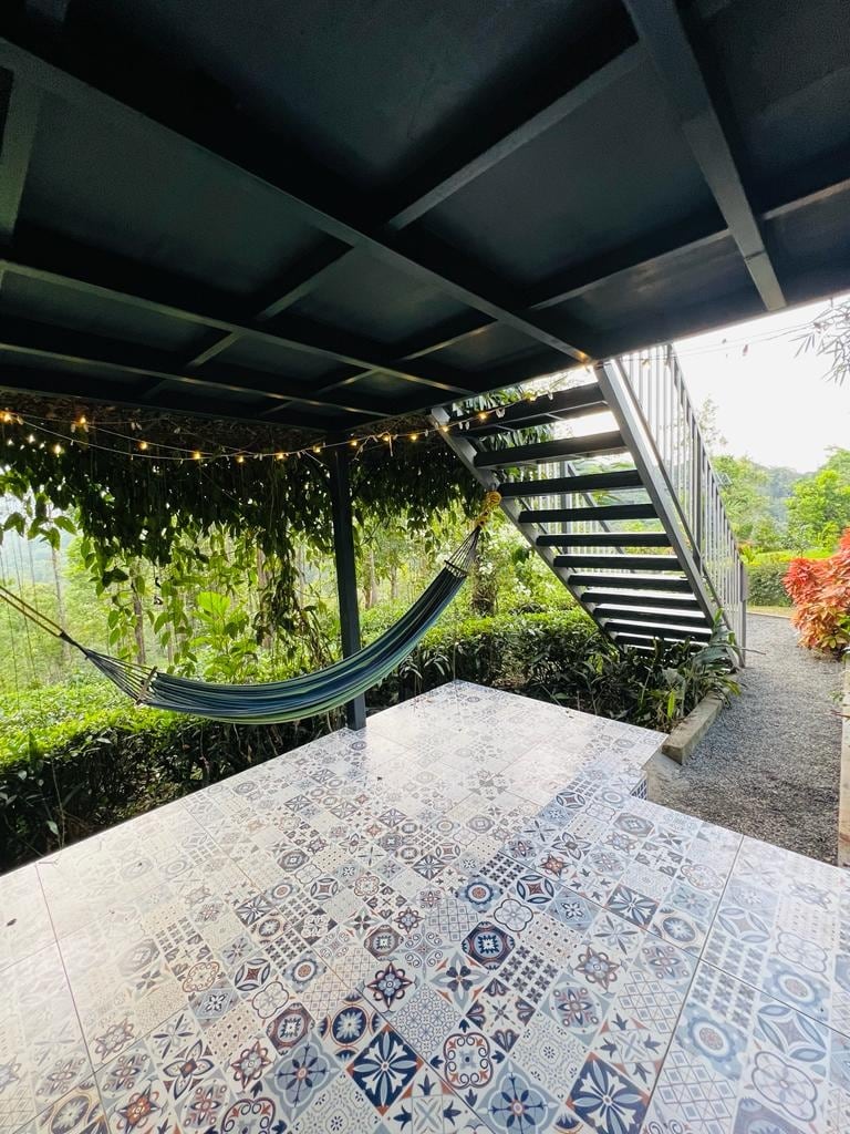 A spacious outdoor area is highlighted by decorative tiled flooring, providing a colorful visual. A hammock is suspended, inviting relaxation. A staircase is visible, leading to an upper level, while surrounding greenery enhances the natural setting and creates a serene atmosphere.