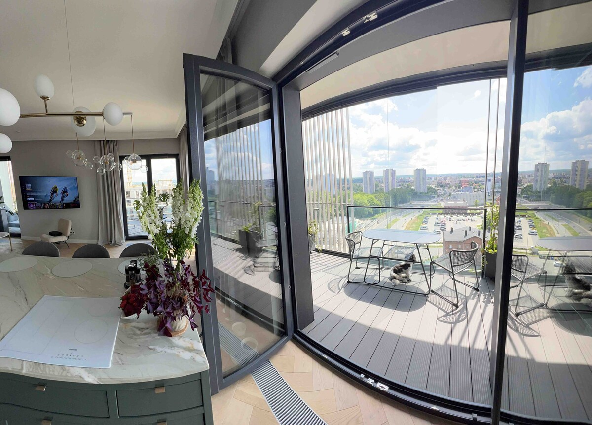 A panoramic view is shown through large glass doors that open to a spacious terrace. Outdoor furniture is arranged for relaxation, surrounded by greenery and a cityscape in the background. Natural light brightens the interior space, enhancing the sense of openness.