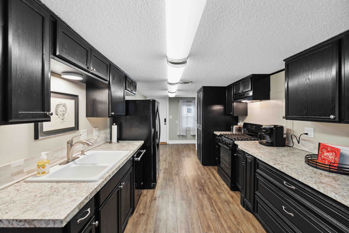 The kitchen offers dark cabinetry paired with light granite countertops, providing a modern aesthetic. An efficient layout includes a double sink, gas stove, refrigerator, and ample storage. Soft lighting enhances the warm tones of the wood flooring, creating a welcoming space for cooking and gathering.