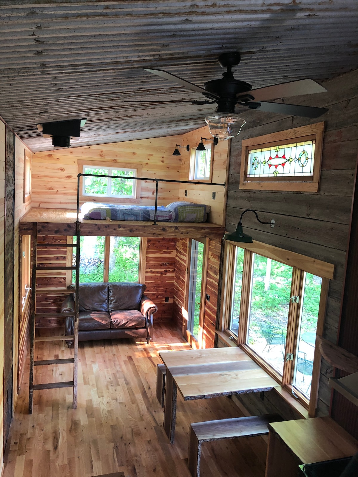 An airy interior showcases wood paneling and a rustic charm. A ladder leads to a lofted sleeping area, with a sturdy couch placed below. A wooden table is positioned near expansive windows, allowing natural light to illuminate the space, complemented by a colorful stained glass feature.