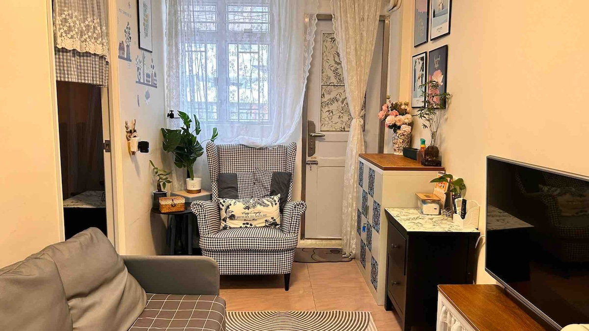 A cozy living area features a checkered armchair beside a door, complemented by light curtains. A small table holds decorative flowers, while a compact cabinet displays various items. A comfortable gray sofa invites relaxation, with a soft area rug underfoot, enhancing the welcoming environment.