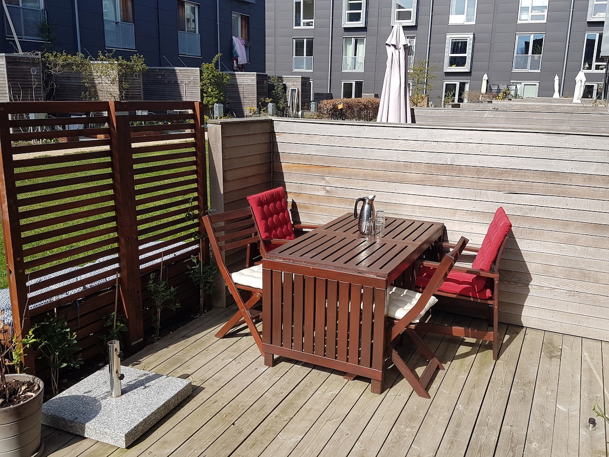 A private terrace is featured with wooden decking, showcasing a rectangular table surrounded by two red cushioned chairs and a matching bench. Planters with greenery add to the space, while a large umbrella is positioned nearby for shade.