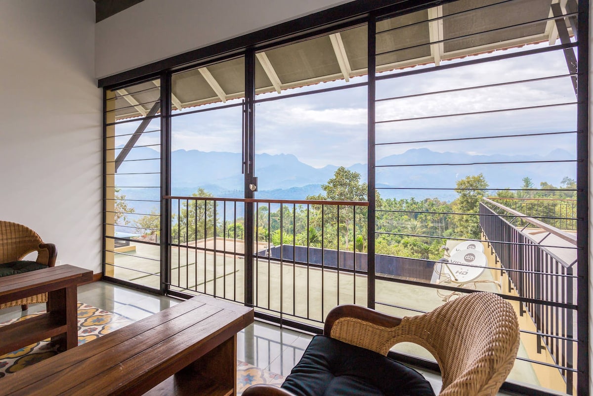 A cozy sitting area features two woven chairs and a wooden table. Large glass doors provide panoramic views of the surrounding mountains and greenery, allowing natural light to fill the space. The balcony enhances the serene atmosphere, inviting relaxation.