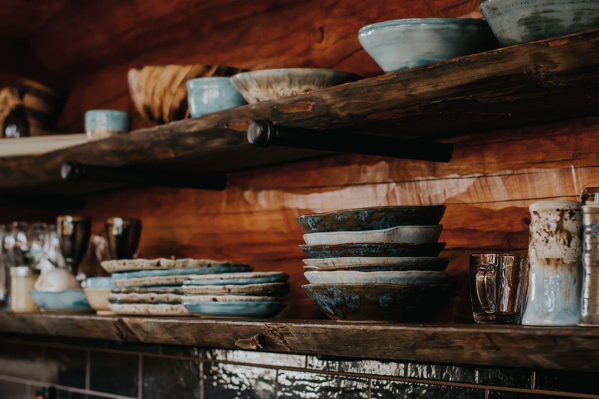 Rustic wooden shelves display a variety of handcrafted pottery, including bowls and plates in soft blue hues. Glassware and metallic cups are neatly arranged alongside, all set against a warm wooden backdrop.