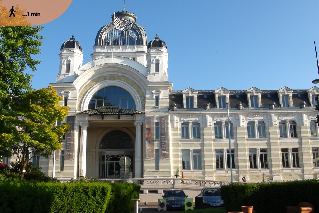 A grand building features an ornate façade with large arched entrances and decorative details. The structure is topped with a dome, and lush greenery lines the perimeter. A small courtyard is visible, complemented by parked vehicles in front of the entrance.