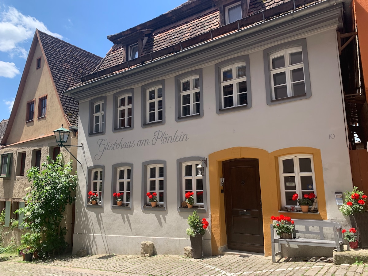 The exterior view of a historic building showcases a charming façade with decorative window boxes filled with red flowers. The entrance features a wooden door framed by warm tones, while multiple windows allow natural light to illuminate the space. A cobblestone pathway leads to the entrance.