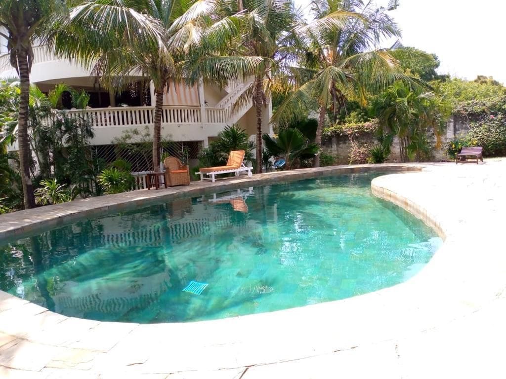 A clean swimming pool is surrounded by palm trees and tropical foliage. Comfortable seating areas are located nearby, offering a relaxing atmosphere. The villa's upper level can be seen in the background, featuring a balcony with simple wooden furniture.