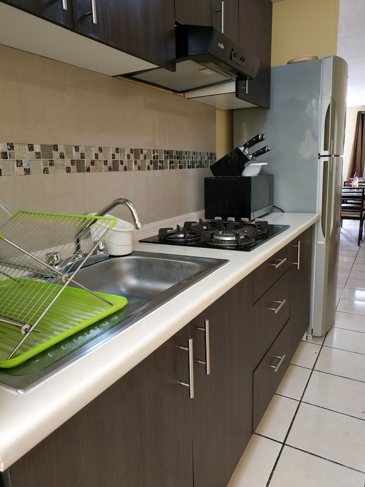 The kitchen area features dark wood cabinetry, complemented by a clean countertop. A stainless steel sink is positioned adjacent to a green dish rack. A gas stove and a refrigerator are included, along with kitchen utensils neatly arranged in a countertop holder.