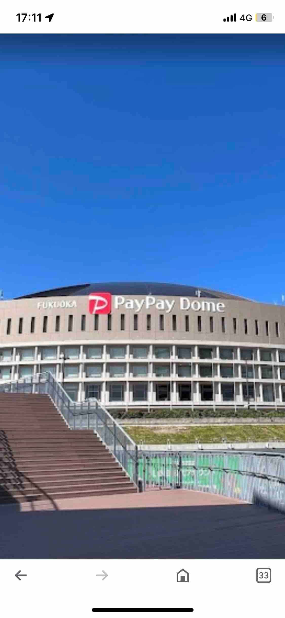 A large stadium is shown featuring a prominent sign that reads 'PayPay Dome.' The building has a modern architectural design with a series of windows and a staircase leading up to it, all set against a clear blue sky.