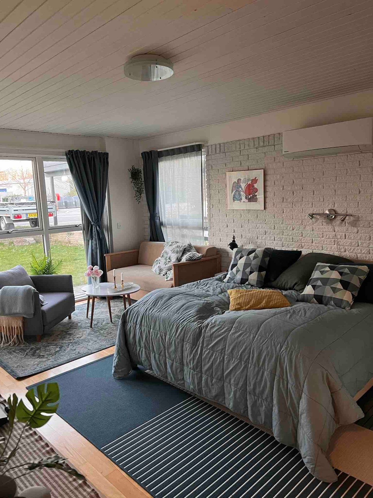 A spacious room contains a comfortable double bed covered with soft bedding, complemented by a sofa and decorative cushions. A small coffee table sits nearby, surrounded by curtains that frame large windows letting in natural light. A rug adds texture to the wooden floor.