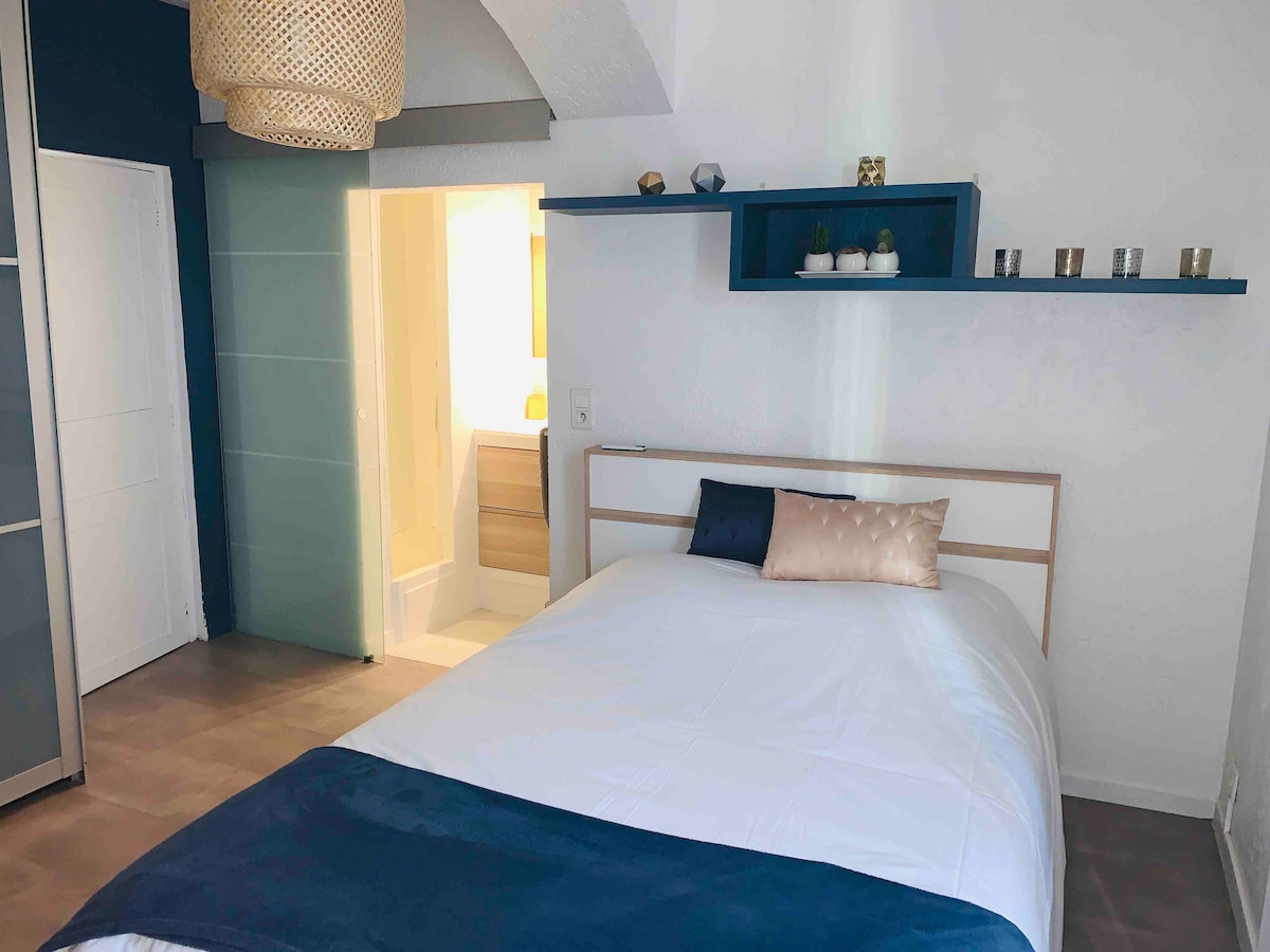 A well-appointed bedroom features a spacious bed dressed in white bedding with a navy throw. A light and airy atmosphere is enhanced by a decorative pendant light. A glass partition offers a glimpse into the adjoining bathroom, while shelves above showcase decorative items.