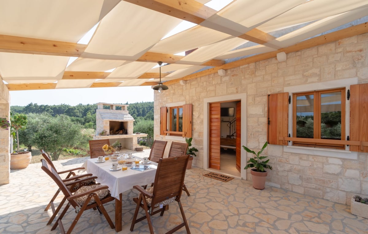 An outdoor dining area is set under a canopy, featuring a stone table laid with breakfast items. Wooden chairs surround the table, and a brick barbecue structure is visible in the background. Olive trees and greenery provide a natural backdrop, enhancing the space's tranquility.
