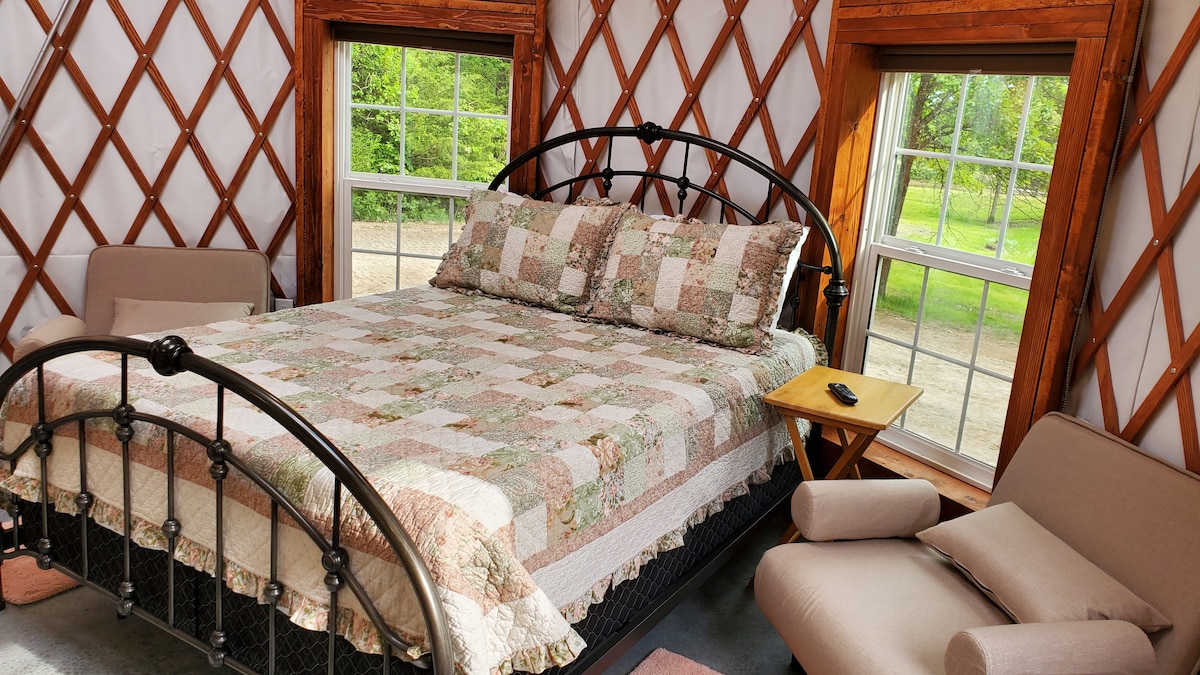 The sleeping area features a spacious bed with a patchwork quilt, complemented by two comfortable chairs. Lattice walls create a unique aesthetic, and large windows provide views of the surrounding greenery, allowing natural light to brighten the space.