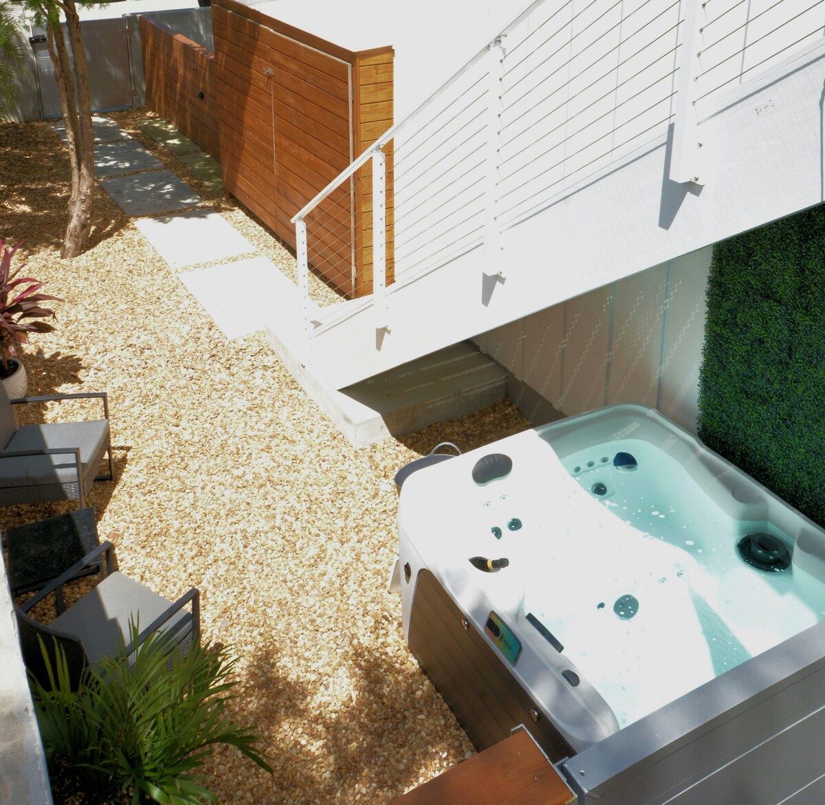 A private courtyard is depicted, featuring a pristine hot tub surrounded by decorative gravel. Comfortable seating is positioned nearby, complemented by lush greenery. Sturdy wooden storage units are visible in the background, along with a stairway leading to the villa’s entrance.