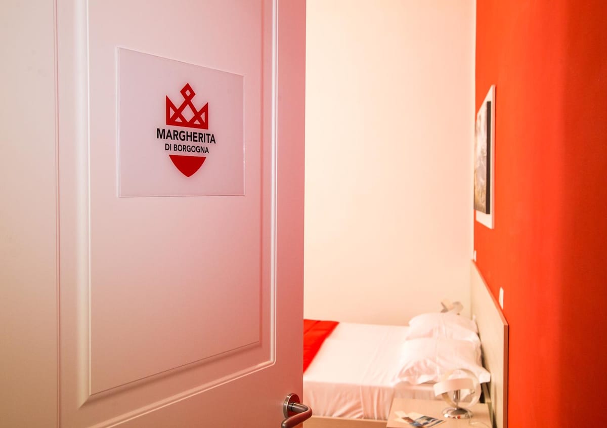 A doorway is shown, revealing a bright room with a warm red accent wall. A spacious bed is made with crisp white linens and a coordinating red throw. Artwork adorns the wall, complementing the modern decor.