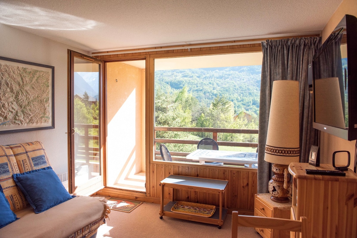 A living area features large sliding glass doors that open to a balcony, allowing ample natural light. Furnishings include a wooden coffee table, a comfortable sofa adorned with blue cushions, and a cabinet. Scenic mountain views are visible from the windows.
