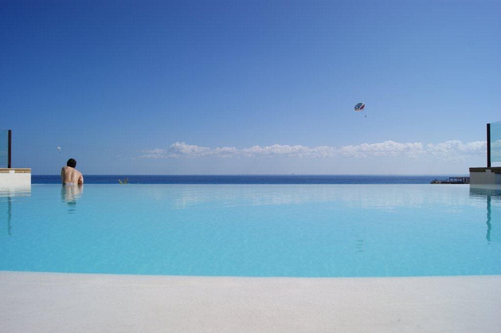 An expansive infinity pool reflects the clear blue sky, seamlessly merging with the horizon. The tranquil water invites relaxation, while a distant view of the ocean highlights the serene setting. A figure is seen enjoying the pool, enhancing the peaceful ambiance.