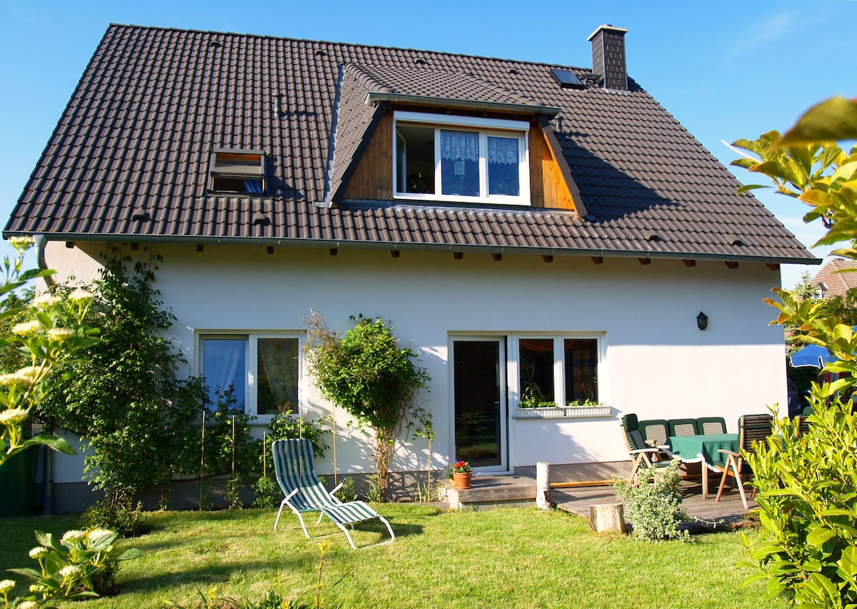 A freestanding house is surrounded by greenery, featuring a sloped roof and large windows. In the foreground, comfortable seating is arranged on a wooden terrace, while a sun lounger rests on lush grass. The overall atmosphere is calm and inviting, perfect for outdoor relaxation.