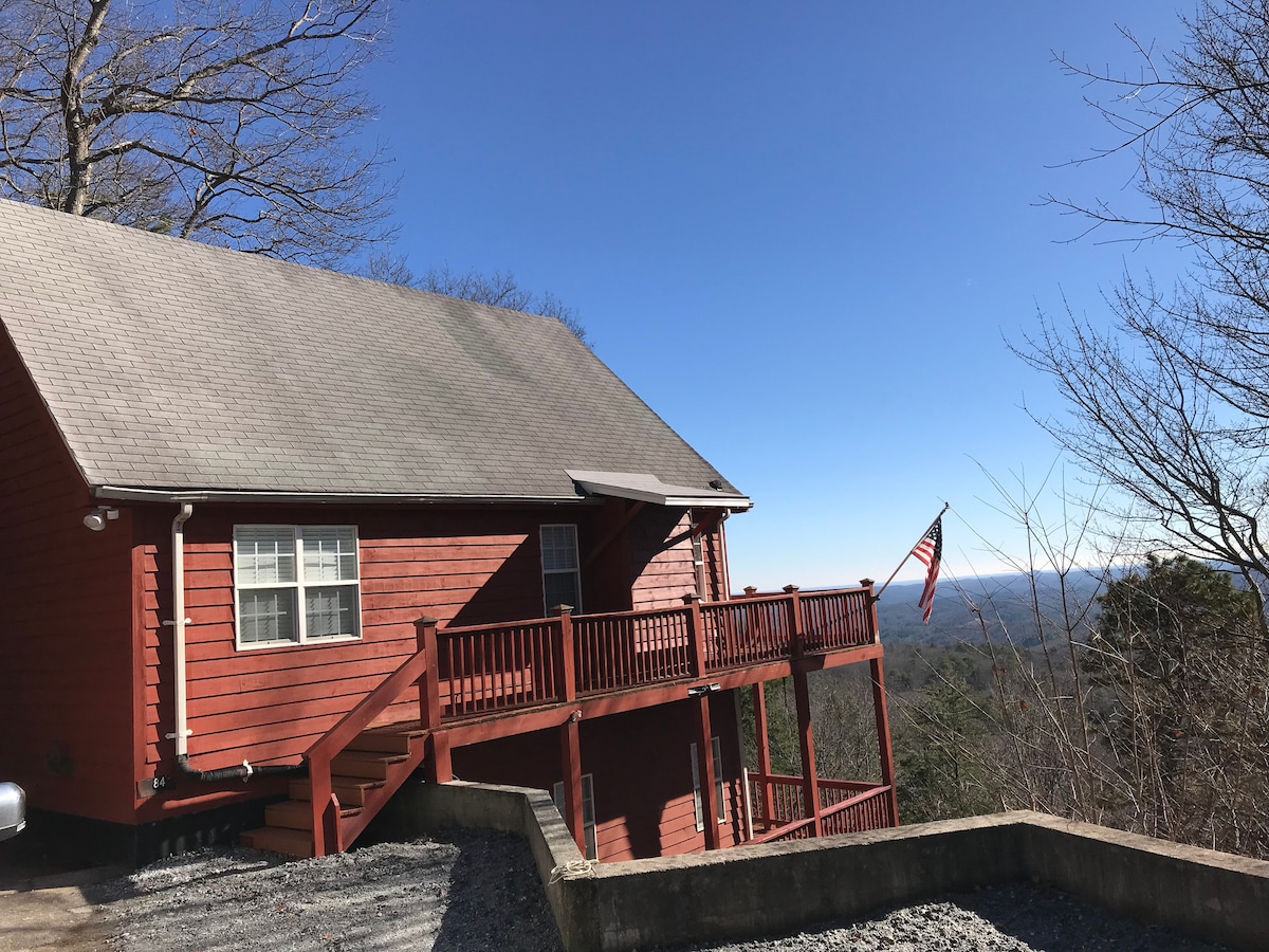 The cabin's exterior features a rich red wood facade, complemented by a wrap-around deck with wooden railing. A flag flutters gently, and panoramic mountain views stretch into the distance under a clear blue sky.