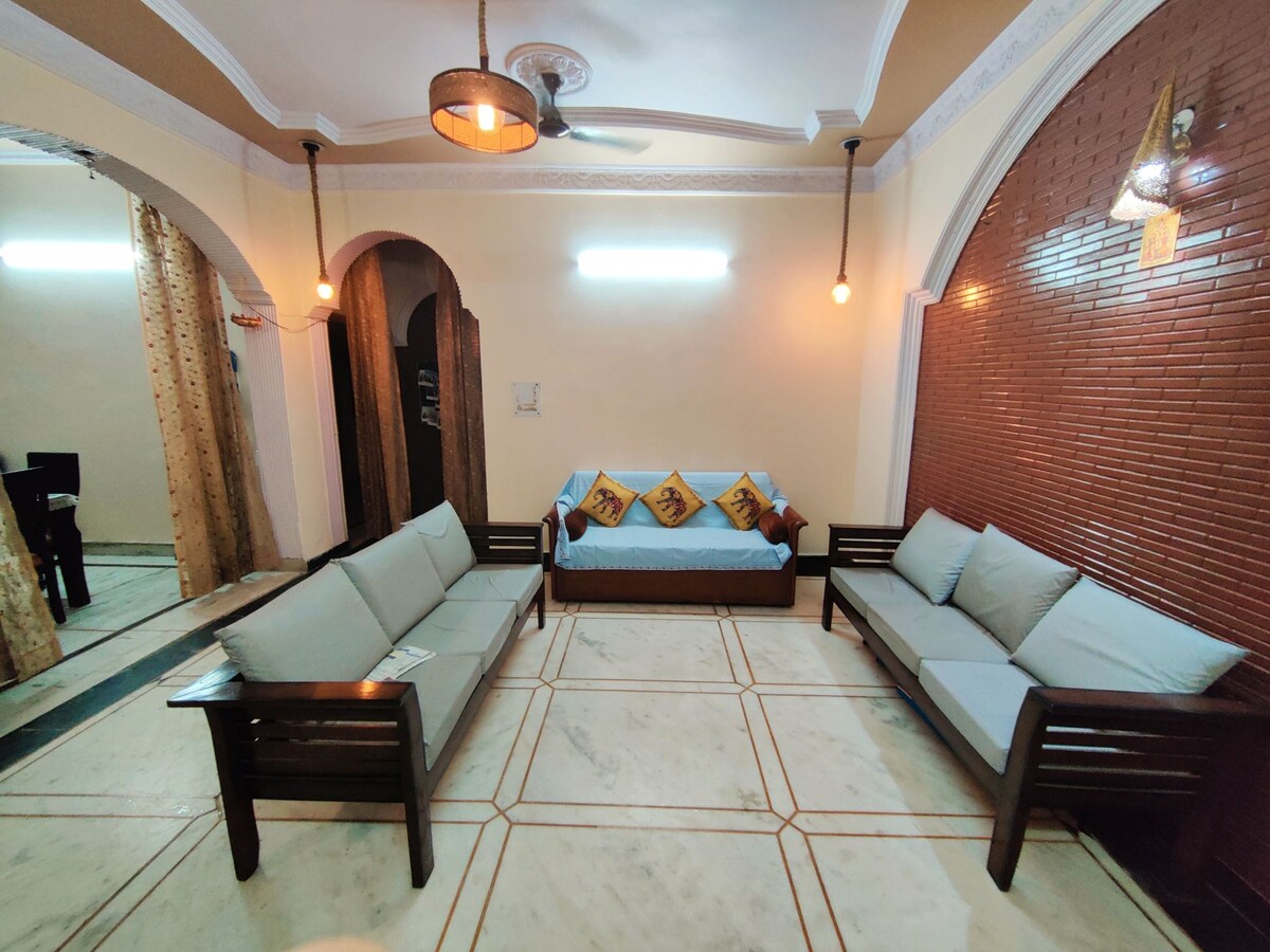 A welcoming seating area features two wooden sofas with light blue cushions positioned around a central space. A sofa bed is placed against the wall, decorated with matching cushions. Soft lighting is provided by hanging fixtures, enhancing the room's comfort.