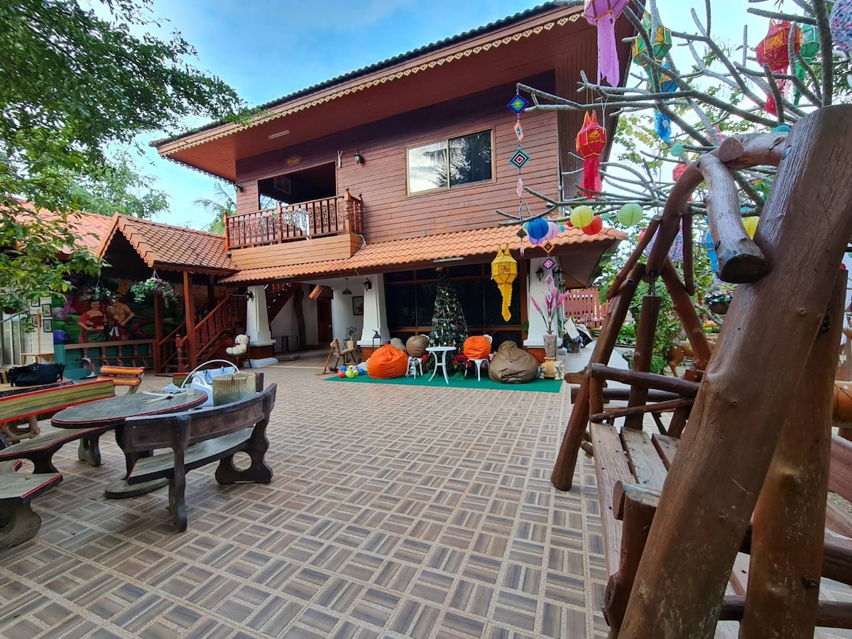 A two-story wooden building is situated amidst lush greenery. The patio area features a mix of playful decorations, including colorful ornaments and various seating options. A swing crafted from natural wood adds to the inviting outdoor space, surrounded by seasonal decorations.