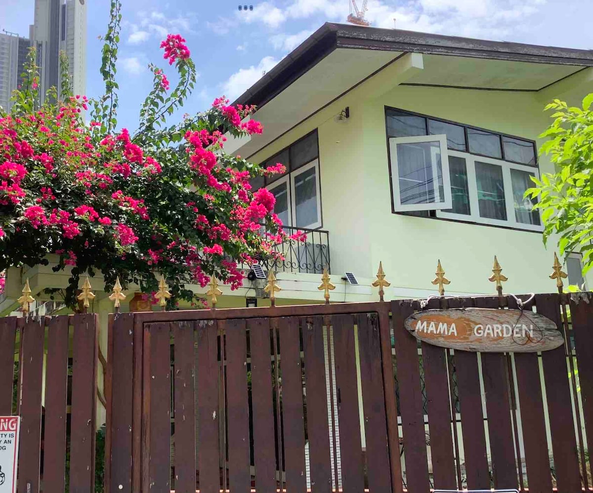 The exterior of a two-story guest house is framed by vibrant pink bougainvillea. A balcony overlooks the entrance, which features a wooden gate adorned with a sign reading 'Mama Garden.' The building is set against a backdrop of cityscape and blue sky.
