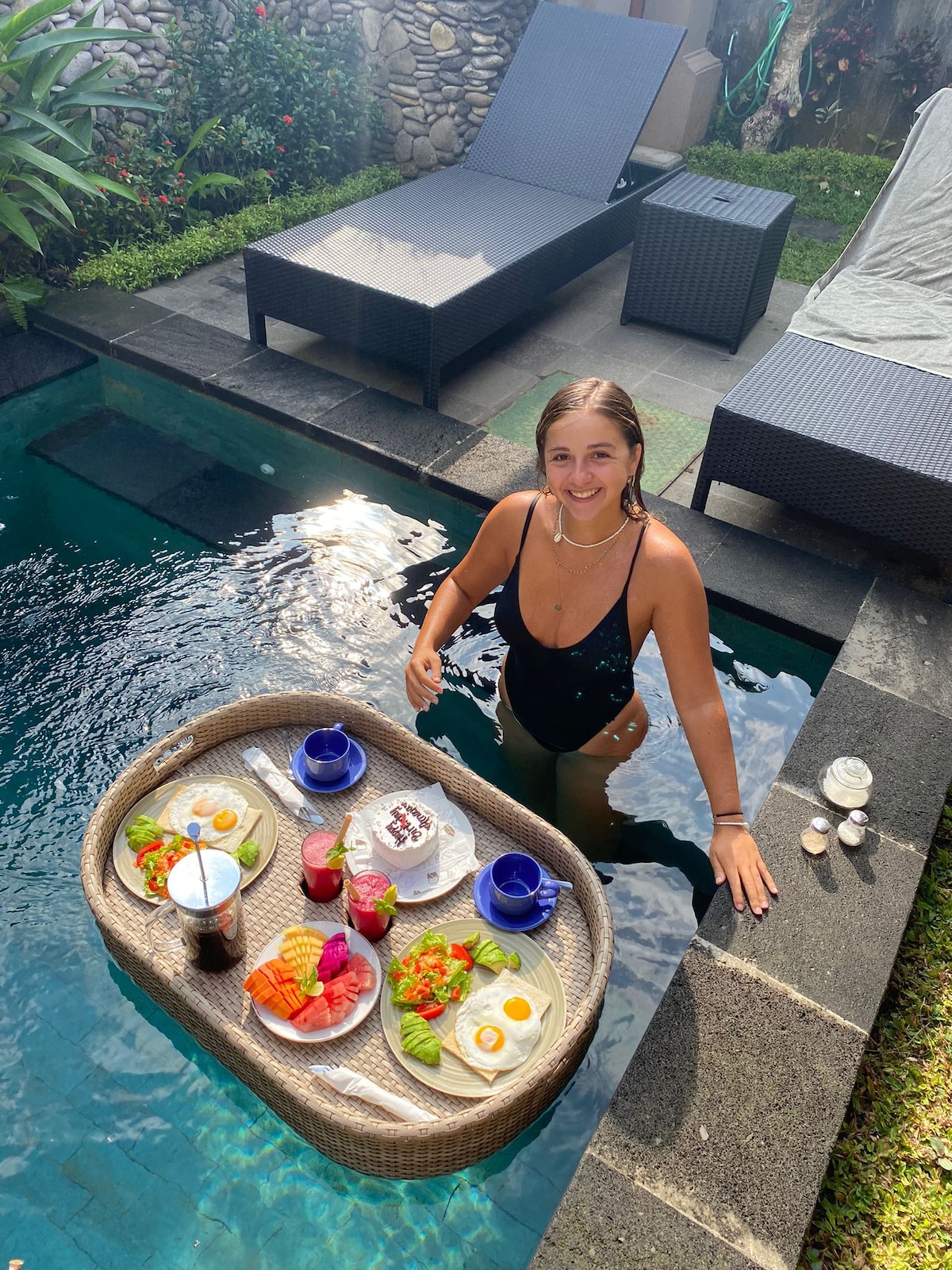 A serene scene features a private outdoor pool surrounded by lush greenery. A floating tray holds an array of vibrant fruit and breakfast items, alongside beverages. Comfortable lounge chairs provide relaxation spots nearby, enhancing the tranquil ambiance of the villa.
