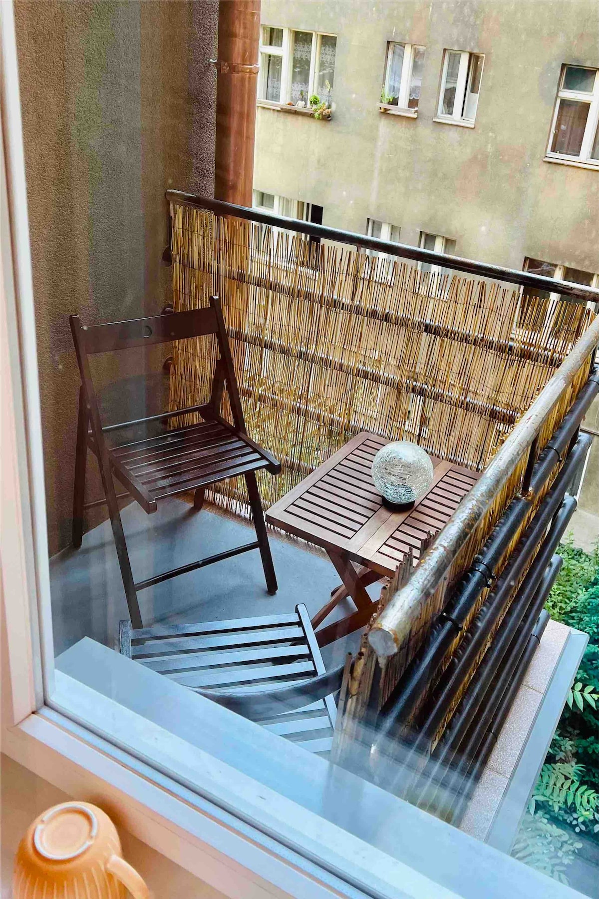 A small balcony is visible, featuring two wooden folding chairs and a square table. A decorative stone sits on the table, surrounded by a bamboo privacy screen. The view overlooks neighboring buildings, and greenery can be seen in the background.