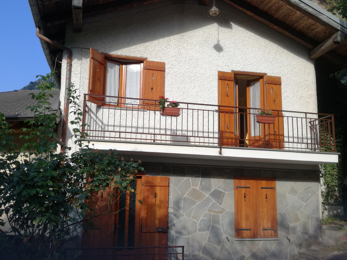 The two-story house features a light-colored facade with wooden shutters. A balcony is visible on the upper floor, adorned with small flower boxes. The stonework at the base complements the overall structure, while greenery surrounds the property, providing a tranquil atmosphere.