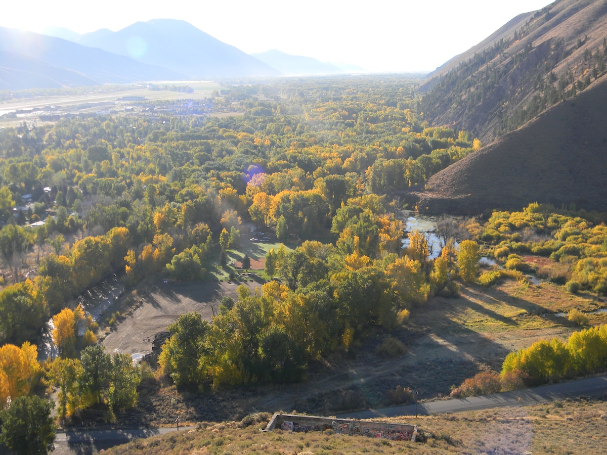 A panoramic view showcases a vibrant landscape with golden autumn foliage covering the trees. The valley stretches into the distance, framed by rolling mountains. The interplay of light and shadow highlights the natural beauty of the area, inviting a sense of tranquility.