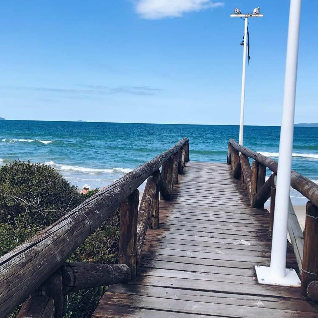 A wooden boardwalk extends toward the ocean, flanked by coastal vegetation. Gentle waves lap against the shore under a clear blue sky, with distant land visible on the horizon. Two light posts stand along the path, providing ambient lighting.