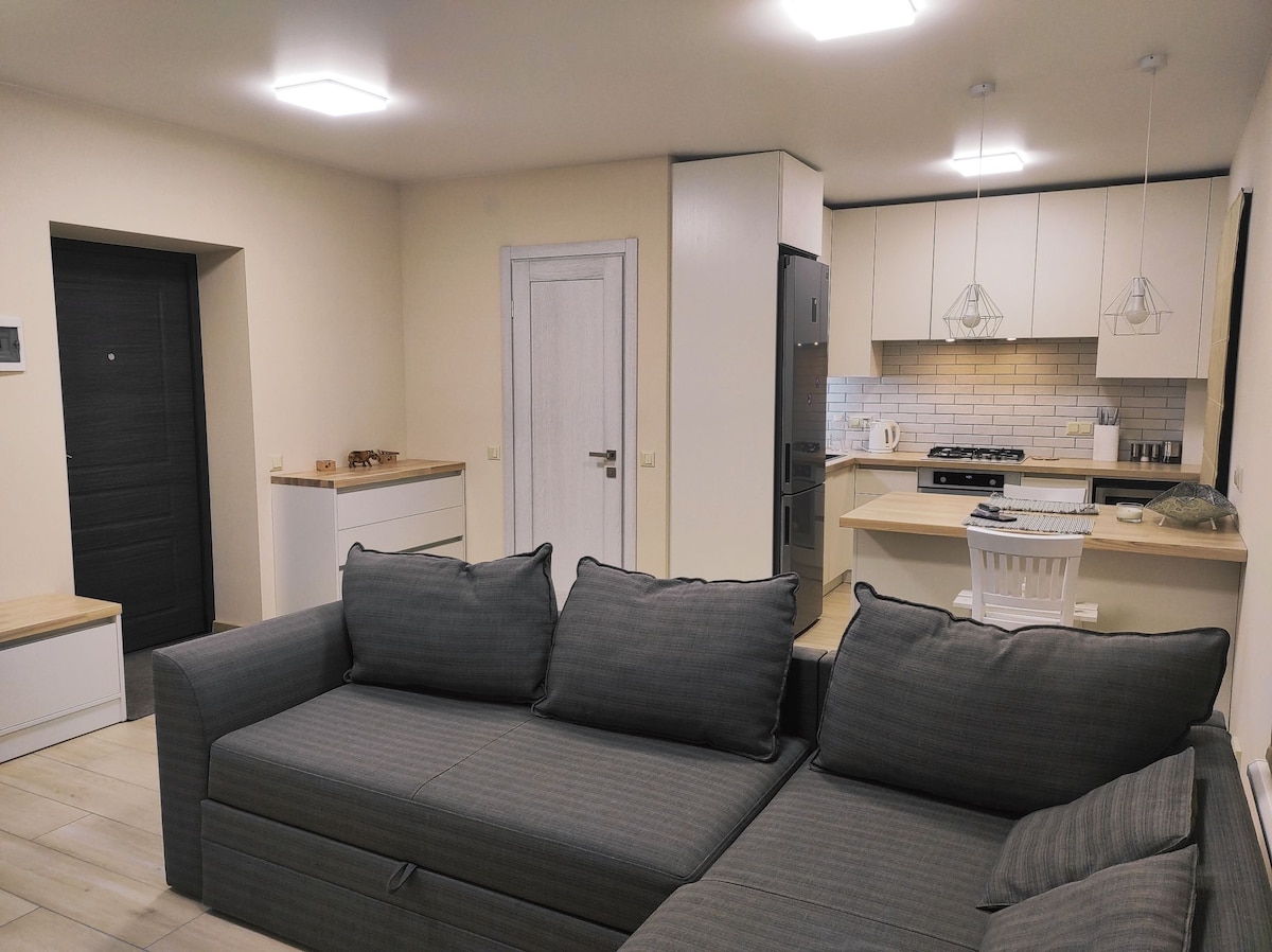 A well-lit living area features a spacious gray sectional sofa with comfortable cushions. In the background, a modern kitchen is visible with white cabinetry and a tiled backsplash. A dining area with a single white chair and light wood accents adds to the cozy ambiance.