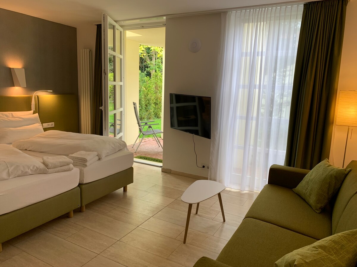A well-lit room features a spacious bed with neatly arranged bedding, alongside a comfortable sofa. Large glass doors offer views of a terrace and garden. A television is mounted on the wall, complementing the room's modern decor and soft, natural colors.